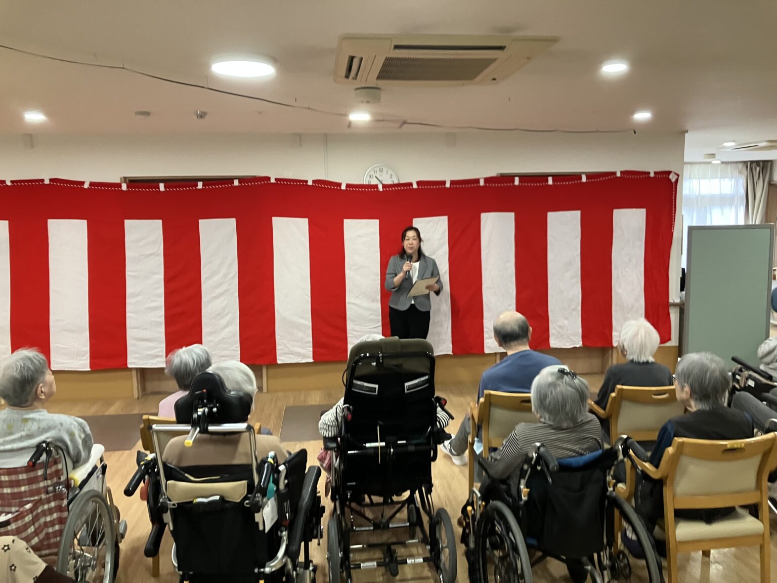 介護施設での催し。赤白の垂幕を背景に、職員が高齢入居者向けに活動プログラムを進行している。